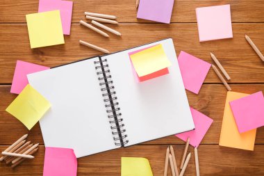 Bir not defteri, çeşitli renklerde parlak yapışkan notlarla çevrili ahşap bir yüzeye uzanır. Kullanılmayan renkli kalemler etrafa saçılmış. Bu da fikir üretimi için düzenli bir çalışma alanı olduğunu gösteriyor..