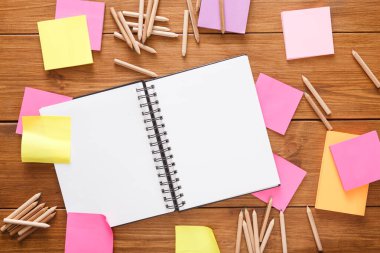 Boş bir spiral defter, pembe ve sarı renkli yapışkan notlarla çevrili ahşap bir yüzeyde yatıyor. Tahta kalemler etrafa saçılmış, yaratıcılığa ve organizasyona davetiye çıkarıyorlar..