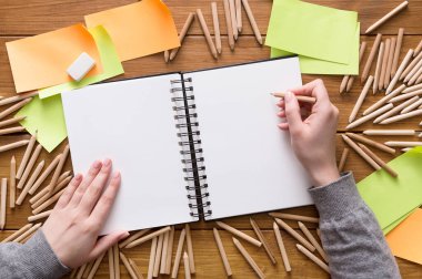 Bir kişi renkli kalemler ve yapışkan notlarla çevrili boş bir deftere çizim yapmaya hazırlanıyor. Ahşap masa çalışma alanına yaratıcı bir atmosfer ekler..