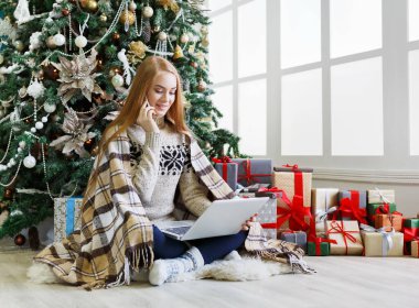 Cep telefonuyla konuşan genç bir kadın ve rahat bir Noel 'de dizüstü bilgisayarla alışveriş yapan bir kadın. Kız süslenmiş çam ağacının altında hediyelerin arasında oturuyor. Xmas 'a hazırlanmak, kış satışlarında hile yapmak, kopyalama alanı