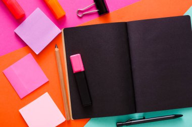 Parlak ve cezbedici çalışma alanı canlı turuncu ve pembe bir yüzey üzerinde açık bir defter içerir. Etrafında yapışkan notlar, bir kalem, bir fosforlu kalem ve bir ataç var..