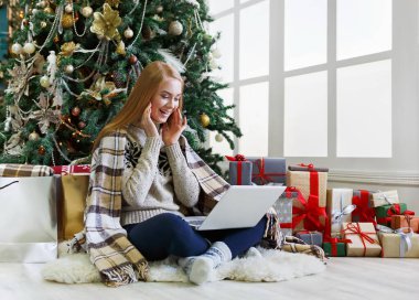 Dizüstü bilgisayarı olan mutlu kız süslü Noel ağacı arka planında oturuyor. Kadın internetten tebrik e-postası almış. Kış indiriminde büyük indirim varmış. Tatil atmosferi, boşluğu kopyala
