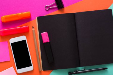 Siyah bir defter, renkli fosforlu kalemler, yapışkan notlar, bir kalem ve bir akıllı telefondan oluşan canlı bir çalışma alanı. Parlak masa arkaplanı ayarlara enerji ekler.