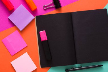 Canlı bir masa düzeneği boş bir siyah defter, renkli yapışkan notlar, bir kalem ve fosforlu kalemler içerir. Parlak arka plan neşeli bir dokunuş ekliyor, çalışmak ya da çalışmak için mükemmel..