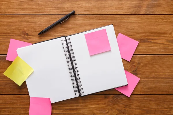 Tahta bir masanın üzerinde açık bir not defteri var. Etrafını renkli yapışkan notlar sarmış. Beyin fırtınası yapıldığını gösteriyor. Not defterinin yanında yazmaya hazır bir kalem var..