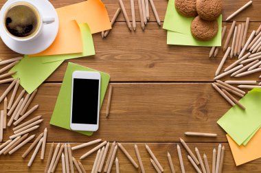 Sıcak bir çalışma odası bir fincan kahve, kurabiye ve renkli yapışkan notlarla düzenlenir. Tahta kalemler akıllı telefonun etrafını sarar ve notlar açar. Fikir üretimi için canlı bir atmosfer yaratır..