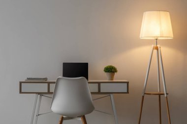 Beyaz koltuk, boş ekranlı dizüstü bilgisayar, masada saksı çiçeği ve akşamları ofis veya oturma odasındaki gri duvarda parlayan lamba. İş yeri, kişisel izolasyon, modern minimalist iç mekan, hiç kimse