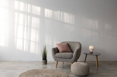 Yastıklı rahat koltuk, masada parlak lamba, saksı çiçeği, kanepe ve yerde yuvarlak halı basit oturma odasındaki gri duvar arka planında. Çağdaş ev tasarımı, İskandinav tarzı.