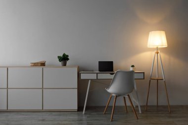 Beyaz sandalye, masadaki boş ekranlı dizüstü bilgisayar, parlak lamba ve akşam çömleğe konan bitki oturma odasındaki gri duvar arka planında. Mobilya reklamı, modern iç mekan hakkında blog, hiç kimse