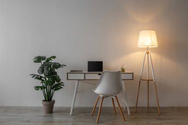 Boş sandalye, boş ekranlı dizüstü bilgisayar, parlayan lamba ve saksı bitkisi akşamları ofis veya oturma odasındaki beyaz duvarda. Modern minimalist iç mekan, işyeri, tasarım reklamı