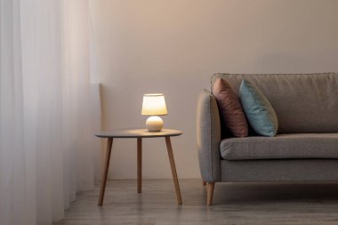 Yastıklı kanepe, gri duvardaki masada parlayan lamba akşam ofiste ya da oturma odasında. Minimalist, modern tarz, İskandinav iç mimarisi ve gayrimenkul satışı ve kiralamayla ilgili blog