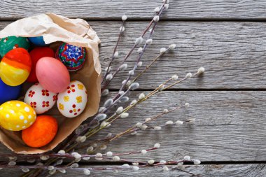 Canlı, süslü yumurtalar kırsal bir masada narin bahar dallarının yanında bir kese kağıdının içinde dinleniyor. Sahne, Paskalya kutlamalarının ve mevsimsel şenliklerin özünü yakalıyor..