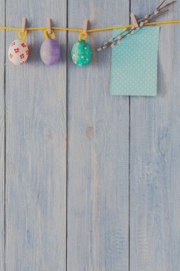 Renkli boyanmış Paskalya yumurtaları mavi ahşap bir duvara tutturulmuş bir ipe asılı. Puantiyeli küçük bir dekoratif not, gösterinin şenlikli atmosferine ekliyor..