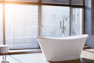 Geniş bir banyoda pencerenin yanında beyaz bir küvet bulunur. Güneş ışığı jaluzilerden geçer, uzayı aydınlatır. Zemin fayanslı ve basit..