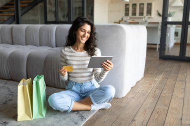 Tabletli bin yıllık beyaz kadın evden alışveriş yaparken kredi kartı kullanıyor, boş alan. Alışveriş torbalarıyla neşeli genç bir kadın uzaktan ödeme yapıyor, web mağazasından mal sipariş ediyor.