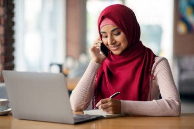 Orta Doğulu olumlu genç bayan işvereniyle telefon görüşmesi yapıyor, rahat bir kafede modern dizüstü bilgisayarın önünde oturuyor, notlar alıyor, internetten iş fırsatları arıyor, yer kopyalıyor.