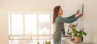 Duvara çerçeve yerleştirirken bir kadın masanın yanında duruyor. Güneş pencereden parlıyor, parlak bir atmosfer yaratıyor. Odada birkaç bitki ve dekor malzemesi var..