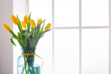 Mavi cam bir vazoda bir buket sarı lale düzenlenir. Vazo bir pencere pervazında duruyor ve içinden doğal ışık geçiyor. Taze yeşillik çiçekleri tamamlar..