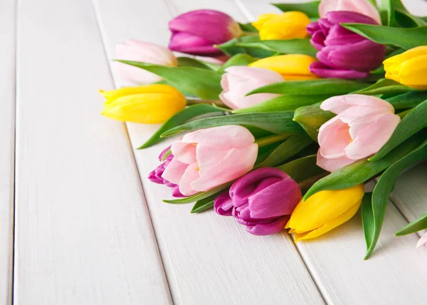 Beyaz ahşap bir yüzeye farklı renklerde taze laleler yerleştirilir. Çiçekler pembe, mor ve sarı tonlarını içerir. Yapraklar yeşil ve çiçekleri tamamlıyor..