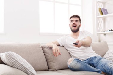 Bir adam, aydınlık bir odada, hafif bir koltuğa rahatça oturur. Uzaktan kumandayı elinde tutuyor ve televizyon ekranına bakıyor. Atmosfer rahat ve kapalı..