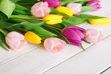 Çeşitli renklerde laleler beyaz ahşap bir masanın üzerine yerleştirilir. Çiçekler taze ve canlı, pembe, sarı ve mor tonlarıyla yeşil yapraklar arasında görünür..