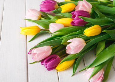 Farklı renklerde laleler açık ahşap bir masada yatıyor. Çiçekler sarı, pembe ve mor çiçekleri içerir. Renkli yaprakları çevreleyen yeşil yapraklar.