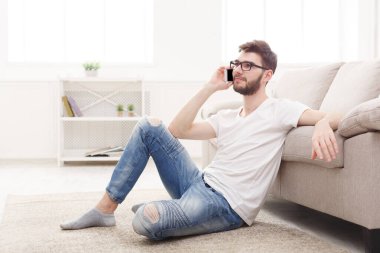 Bir adam kanepeye yaslanmış, yerde oturuyor. İyi aydınlatılmış bir odada telefonla konuşuyor. Mekanın sade bir tasarımı var, rafı ve halısı var..