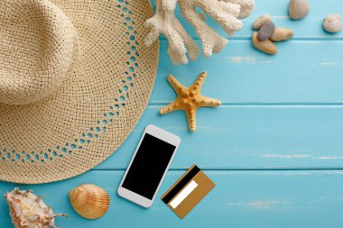 Mavi ahşap üzerinde tatil arka planı, fotokopi alanı olan üst manzara. Plaj aksesuarları cep telefonu, hasır şapka, kredi kartı ve deniz kabukları.