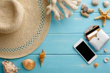 Mavi ahşap üzerinde tatil arka planı, fotokopi alanı olan üst manzara. Plaj aksesuarları- cep telefonu, hasır şapka, kredi kartı ve deniz kabukları.