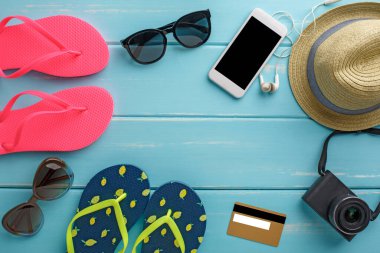 Mavi ahşap üzerinde tatil arka planı, fotokopi alanı olan üst manzara. Plaj aksesuarları, parmak arası terlikler, şapka, kredi kartı kamerası ve boş akıllı telefon.