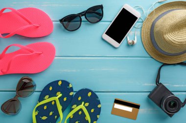 Mavi ahşap üzerinde tatil arka planı, fotokopi alanı olan üst manzara. Plaj aksesuarları, parmak arası terlikler, şapka, kredi kartı kamerası ve boş akıllı telefon.
