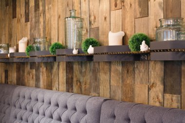 Modern iç mekanlarda dekoratif raflarla süslenmiş ahşap bir duvar sergileniyor. Her rafta cam kavanozlar, beyaz seramik parçalar ve canlı yeşillikler var. Lüks gri koltuklar uzaya rahatlık katıyor..