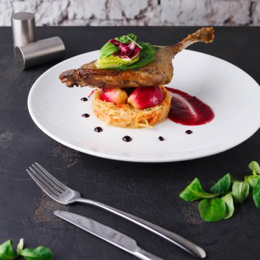 Bir ördek budu tabağı sebze servisinin tepesinde oturur. Tabakta kırmızı sos ve bir garnitür yeşillik var. Dekor, yanında çatal bıçak takımı olan siyah bir yüzey gösteriyor..