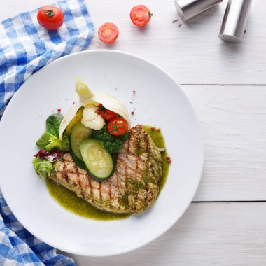 Izgara balık filetosu beyaz bir tabakta servis edilir. Ona pişmiş sebzeler ve yeşil bir sos eşlik eder. Masanın yanında kiraz domatesleri ve mavi kareli kumaş var..