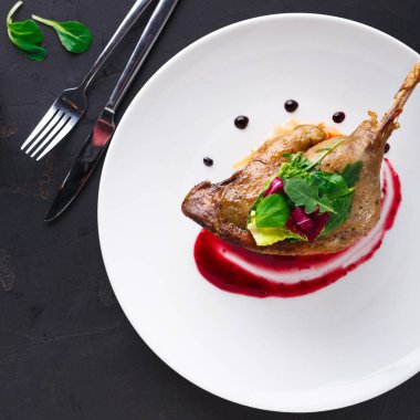 Tabakta kırmızı soslu ve taze yeşilliklerle süslenmiş beyaz bir tabakta ördek bacağı yer alıyor. Tabağın yanına bir çatal ve bıçak yerleştirilmiş. Bu da restoranda yemek yediğini gösteriyor..