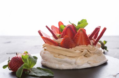Bir pavlova tatlısının üstünde krem şanti ve dilimlenmiş çilek olan siyah bir tabak vardır. Tatlının etrafında taze çilek ve nane yaprakları var. Arka plan parlak..