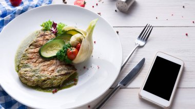 Karışık sebzeli ızgara balık tabağı beyaz bir tabağa konur. Ahşap bir masada tabağın yanında bir akıllı telefon yatıyor. Ayarlar gündelik ve akşam yemeği için uygun..