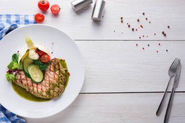 Yanında brokoli, karnabahar ve dilimlenmiş domatesle ızgara balık olan bir tabak. Yeşil sos tat katar. Tabak yakınındaki ahşap bir masada duruyor..