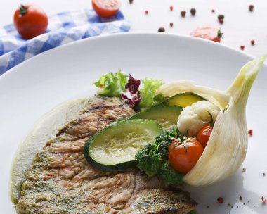 Izgara bir et parçası beyaz bir tabağa konur. Tabakta dilimlenmiş kabak, kiraz domatesleri, brokoli ve sebze karışımı yer alıyor. Yakınlarda mavi beyaz bir kumaş var..