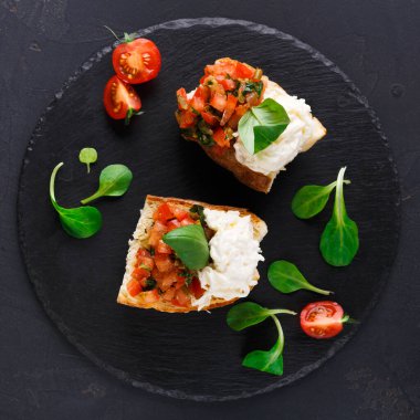 İki parça bruschetta siyah bir tabağa konur. Birinde domates karışımı ve fesleğen, diğerinde ise peynir var. Bruschetta 'yı çevreleyen kiraz domatesleri ve yeşiller.