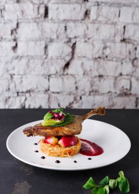 Bir ördek bacağı, beyaz bir tabakta sebze ve meyve yatağının üzerinde duruyor. Tabakta modern bir restoranda tuğla bir duvara dayanmış renkli süsler ve bir sos yer alıyor..