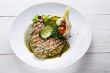 Bir tabak, dilimlenmiş salatalık, kiraz domatesleri ve rezene yanında ızgara balık içerir. Tabak yeşilliklerle süslenir ve ahşap bir masaya yerleştirilir..