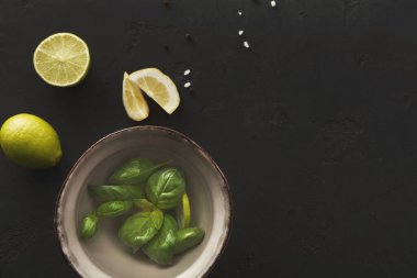 Bir kase suya batırılmış taze fesleğen yaprakları içerir. Yakınlarda, dilimlenmiş limon ve kireç karanlık bir yüzeye yerleştirilir. Bu ayar bir pişirme işlemi için hazırlık olduğunu gösterir.