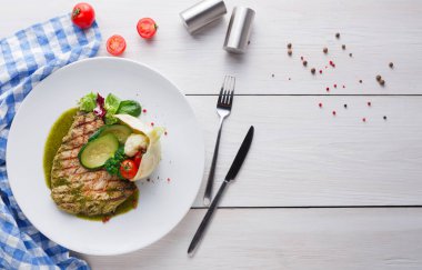 Izgara tavuklu bir tabak açık ahşap bir masada oturur. Tabakta sos, sebze ve domates yer alıyor. Yakınlarda bir çatal ve bıçak var. Baharatlar tabağın etrafına saçılmış..