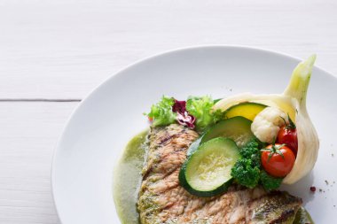 Tabakta açık yeşil soslu ızgara balık filetosu var. Taze sebzeler arasında salatalık, brokoli, karnabahar, marul ve balığın etrafına düzgünce dizilmiş domatesler yer alıyor..