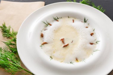 Bir kase kremalı çorba üzerine kızarmış ekmek ve taze otlar koyar. Tabak, yanında yeşil otlar ve basit bir fon bulunan bir masanın üzerine kurulmuş. Bu sahne yemek sunumuna odaklanmış durumda..