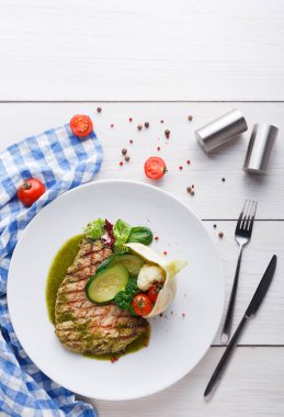 Salatalık ve vişneli domates gibi taze sebzelerin yanında ızgara somon bulunur. Masada mavi kareli peçete, gümüş tuz ve biberlik var..