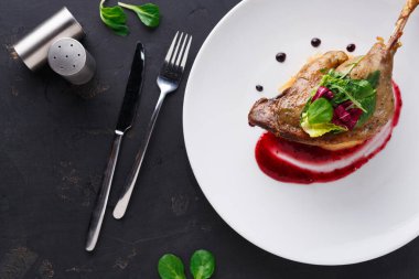 Bir tabak, yeşillik ve kırmızı sosla süslenmiş bir ördek tabağı tutar. Tabağın yanında bir çatal ve bıçak var. Yakınlarda bir tuzluk var. Yemek için ortam hazır görünüyor..