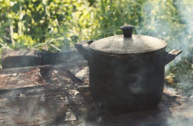 Bir tencere, taş ve metalden yapılmış bir sobanın üzerinde duruyor. Yemek pişirirken çömlekten duman yükselir. Arka planda yeşil bitkiler görülebilir. Gündüz oldu ve güneş parlıyor..