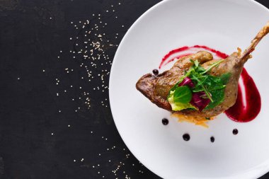 Tabakta pişirilmiş ve süslenmiş bir ördek bacağı var. Etrafı sos ve taze yeşilliklerle çevrili. Karanlık bir arka plan ve dağınık malzemelerle ortam çok basit..